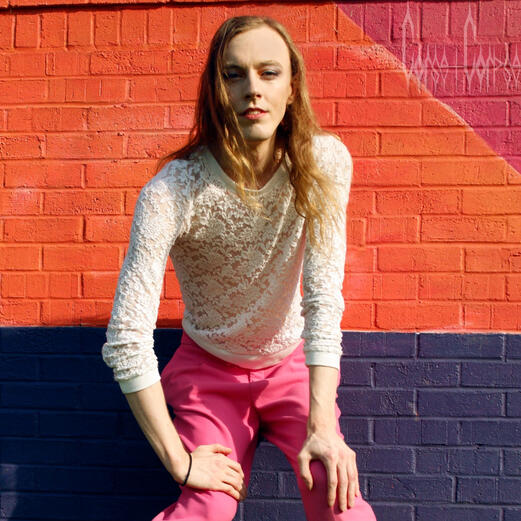 Alaaniel in a white lace, long sleeved shirt and hot pink slacks half-sits against a wall painted orange, blue, and red, with hands on knees and face smoldering forward